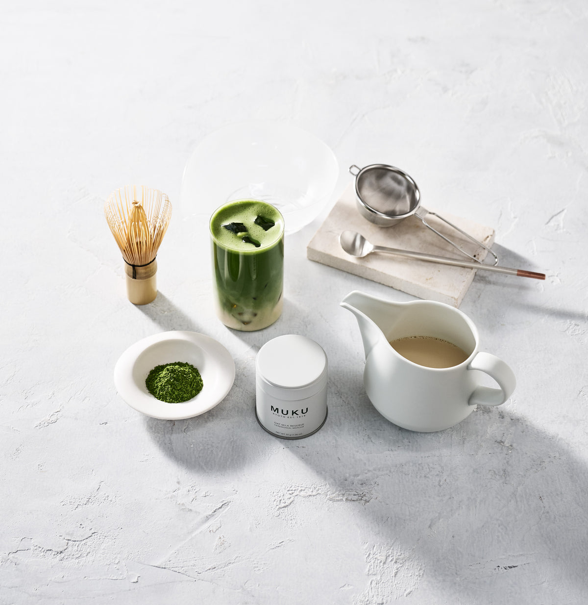 A MUKU matcha latte with a MUKU tin, matcha powder, a bamboo whisk, a sifter and a pitcher of oat milk