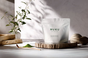 MUKU Oat Milk Reserve 60g pouch on a wooden board beside matcha powder and an olive branch